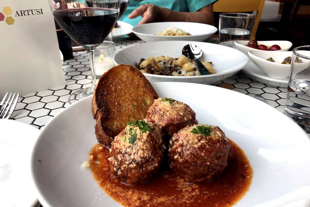 Table level view of several dishes: meatballs and bread with marinara, pasta and mushrooms, and sides with two glasses of wine and water at Pasta night at Artusi, a delicious cheap date idea in Seattle.