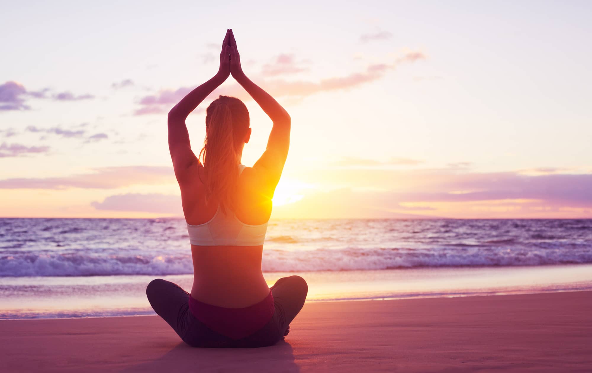 Woman sitting on the sand on a beach doing a yoga pose while the sun sets over the ocean