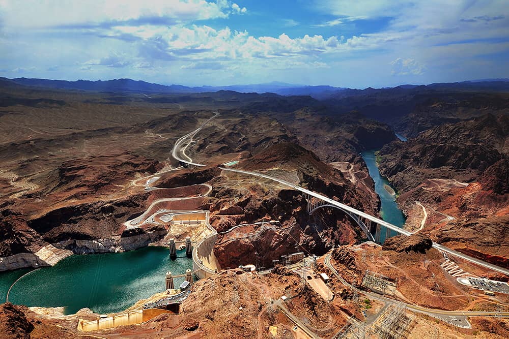 Aerial view of the Mike O’Callaghan-Pat Tillman Memorial Bridge, Hoover Dam, and the Colorado River surrounded by rugged rock formations and a slightly cloudy blue sky