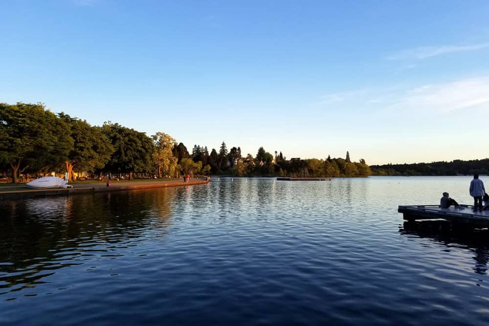  A couple on a dock beside Green Lake on a still, sunny day. The lake is surrounded by a walking path and many green trees, making it a lovely cheap date idea in Seattle