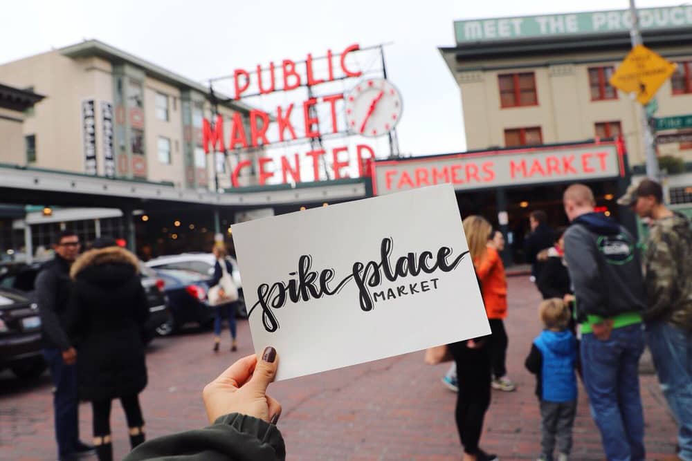 Hand holding a card that reads "Pike Place Market" in front of the farmers market entrance at one of the best cheap date ideas in Seattle