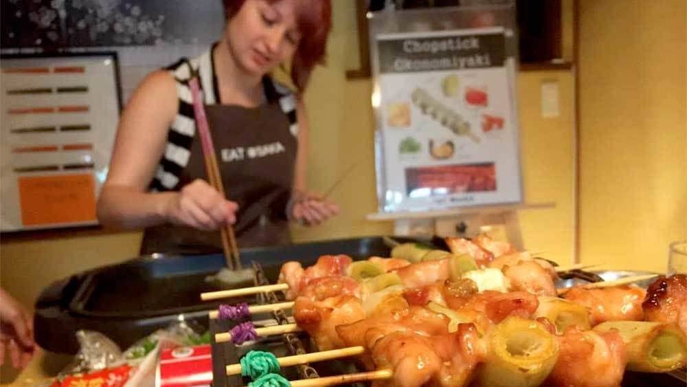 A woman wearing an apron grilling food using long chopsticks while glazed kabobs heat on a barbeque