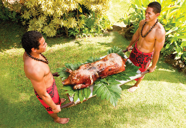 Two men in traditional clothing hold onto roasted pig on tray in Honolulu
