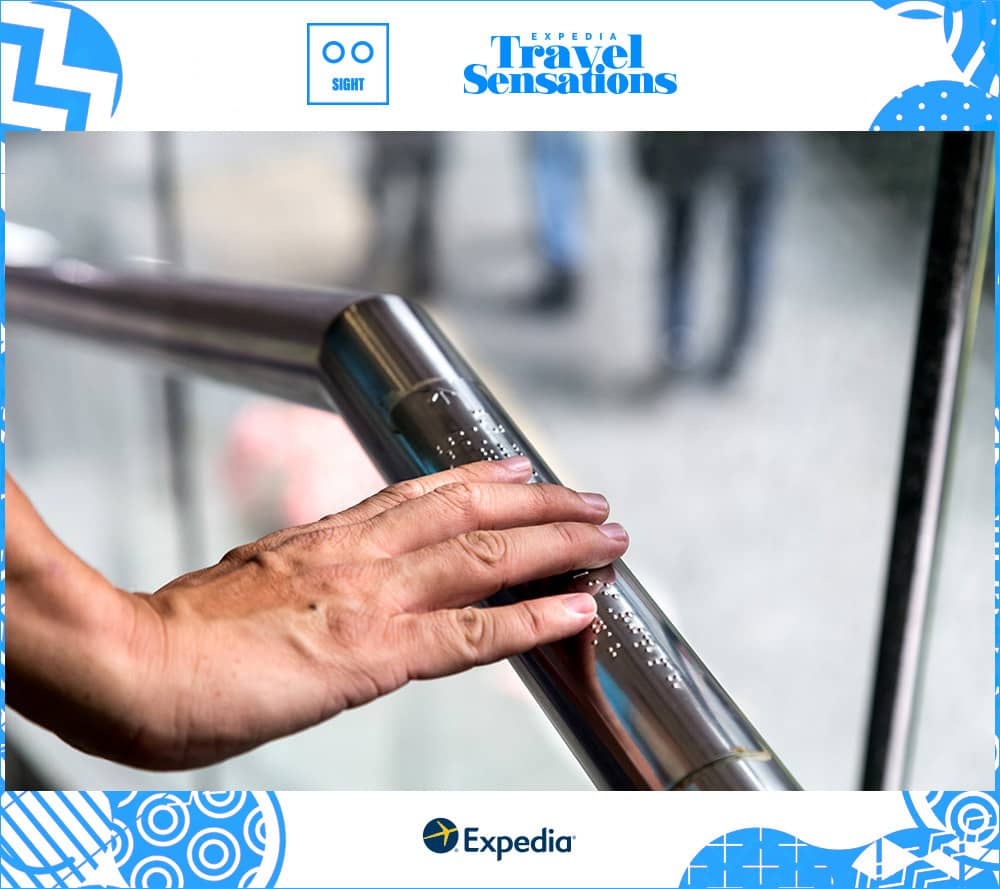 Traveler's hand reads the braille on a metal handrail in front of a window.