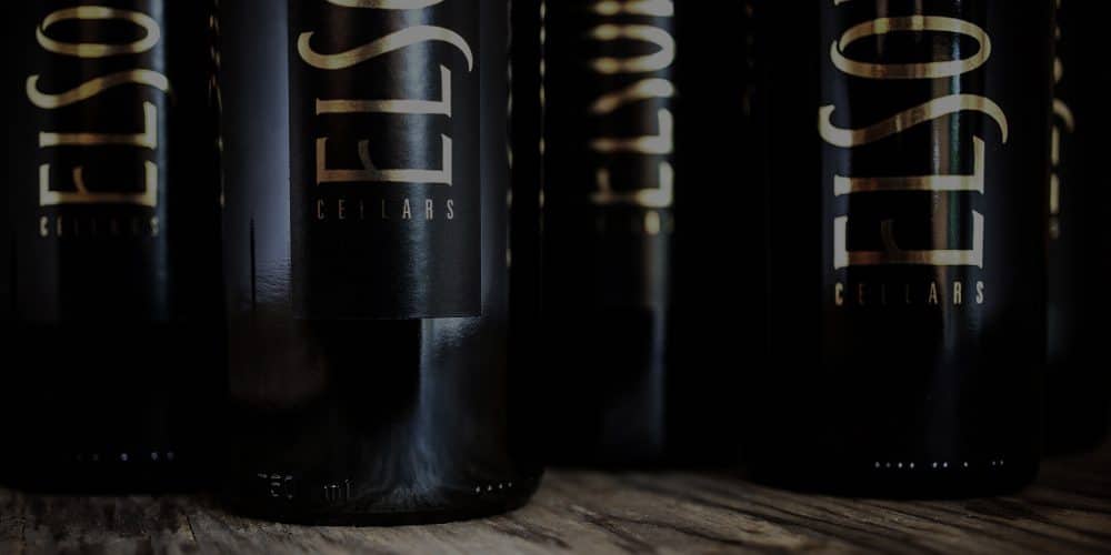 Elsom Cellars wine bottles are black with gold lettering and there is a row of these wine bottles on top of a wooden table surface.