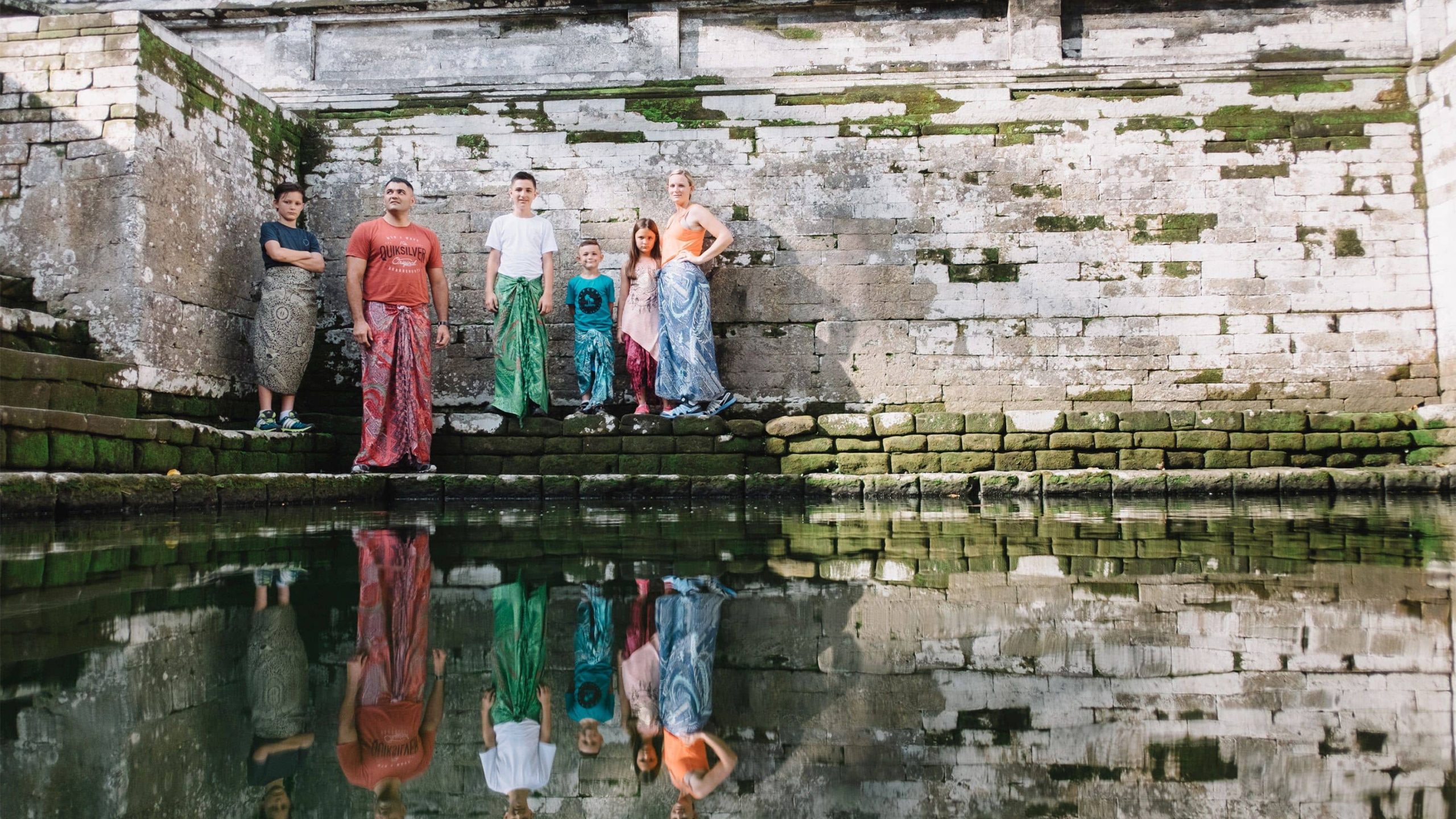A family of six wearing batik print clothing posing for a photographer in front of a reflective pond in Bali