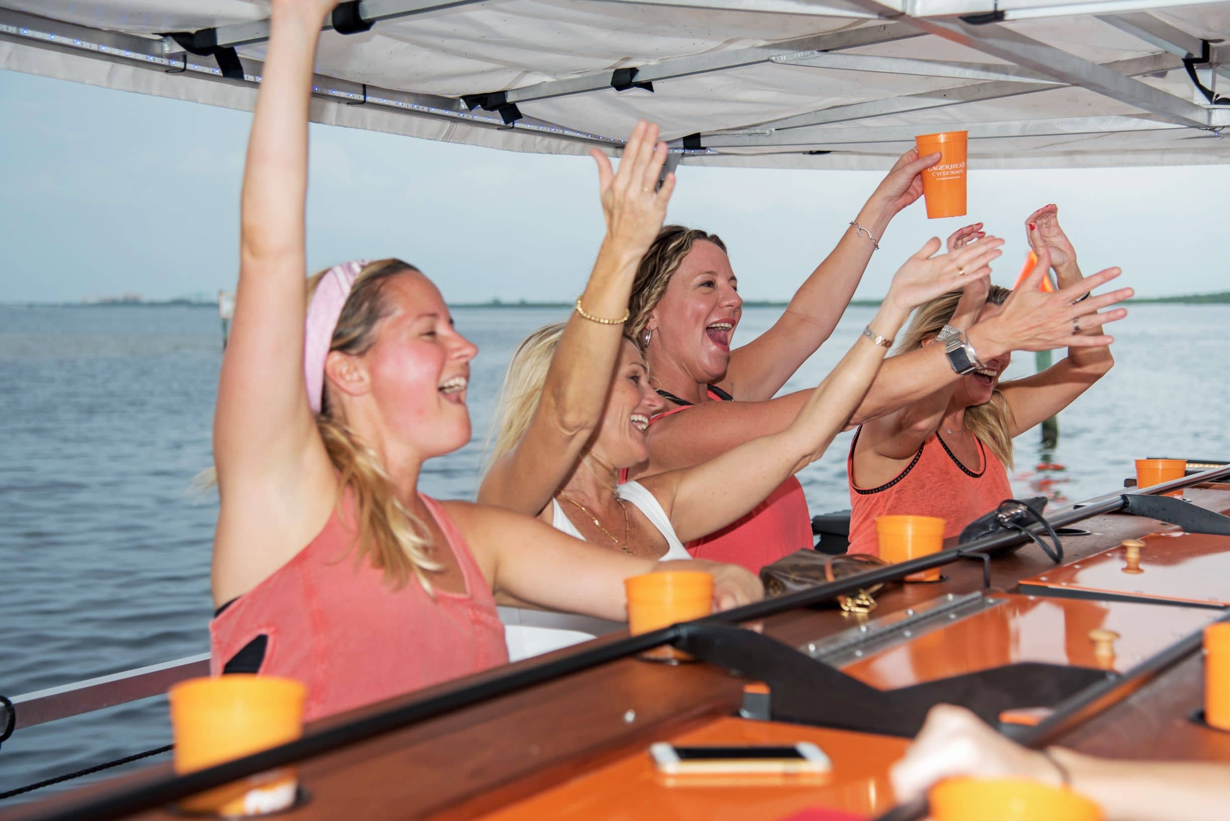 Friends raise their hands and cups in the air aboard a party boat, cheering.