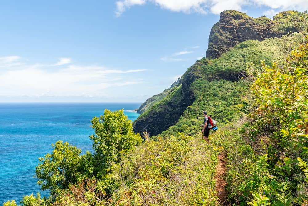 A man hiking a beautiful coastal trail with lush green valleys and cliffs along a bright blue ocean view while traveling solo in Kauai, Hawaii.