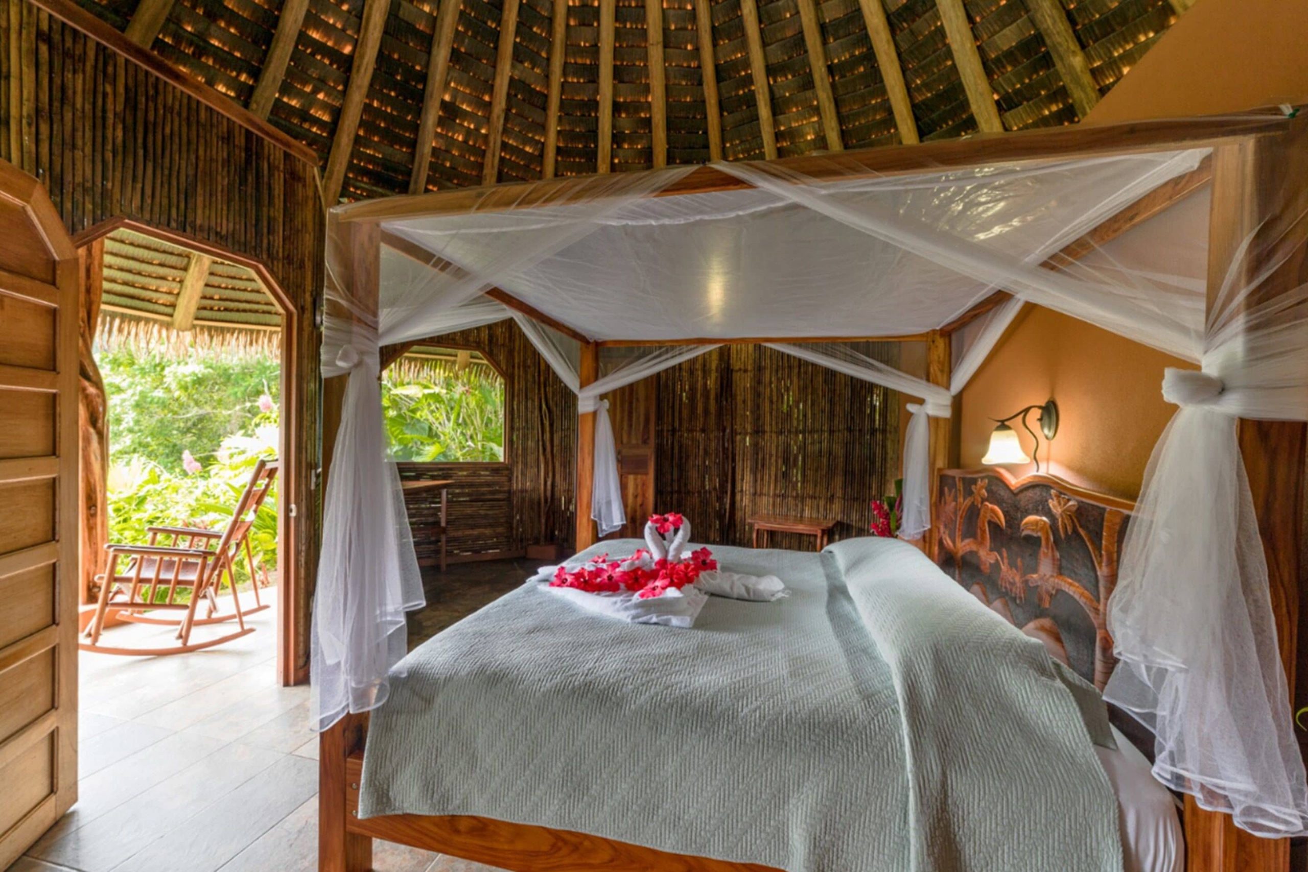 One of the villas for guests to stay in at Luna Lodge in Costa Rica with a four post bed, canopy, white linens, beautiful wooden decor, and an outdoor patio with rocking chairs.