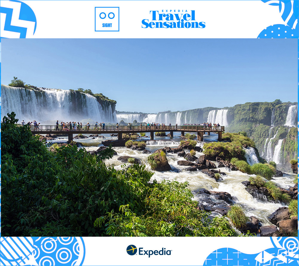 A long bridge curves over the rocks and river currents in front of the curtain-like Iguazu Falls with lush trees and plants growing on the cliffs and rock formations.