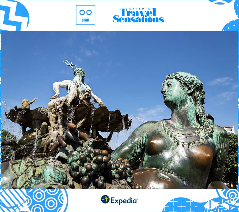  In the foreground a metal figure of a woman with wavy hair and bare skin holds a bunch of grapes, and Neptune sits atop the fountain's upper basin with his trident raised to the clear blue sky.