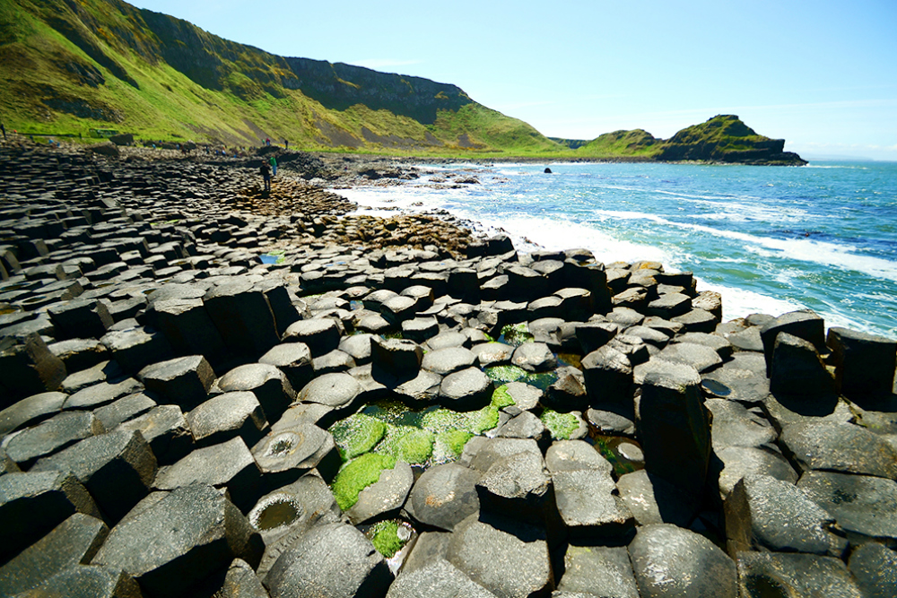 Wet black and grey hexagonal-shaped stones along the coastline, with green mountain slopes in the background at Giant’s Causeway. 