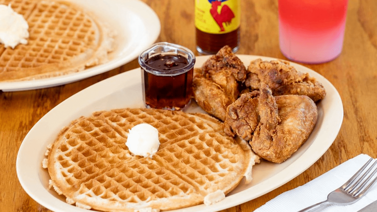 A meal served at Roscoe's Chicken & Waffles in Los Angeles, CA with a large waffle, butter, maple syrup, and fried chicken, all available at cheap prices for big portions.