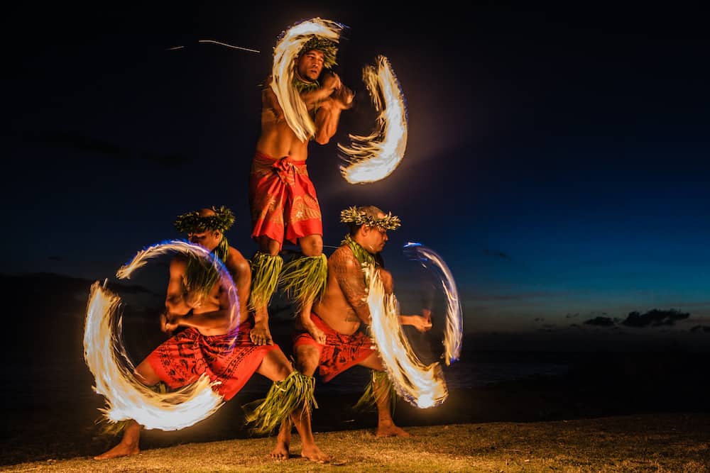 Three men in traditional Hawaiian dress juggle fire on the beach at night.