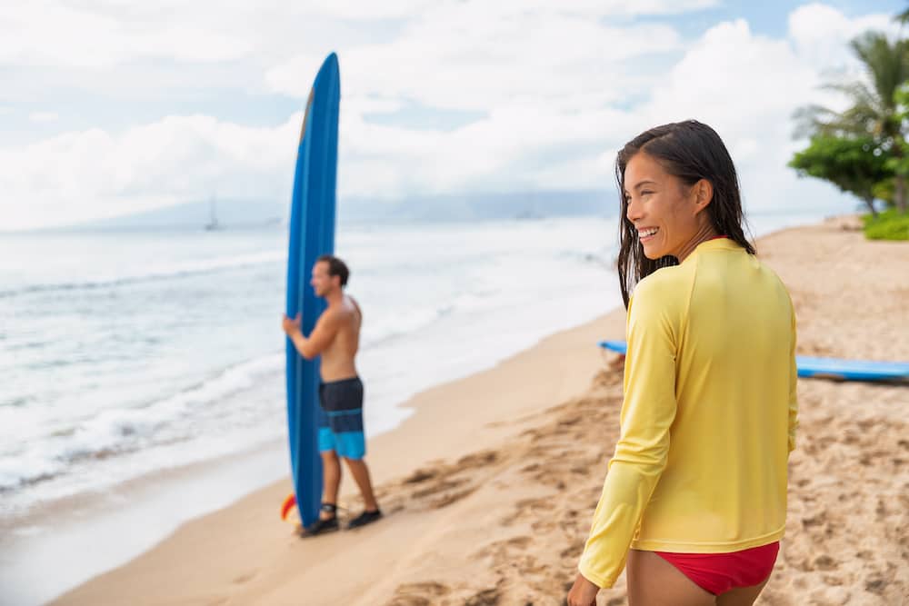 A woman is standing on the beach wearing a rash guard top and looking out at the waves. She is participating in surfing lessons while on a solo trip to Hawaii.