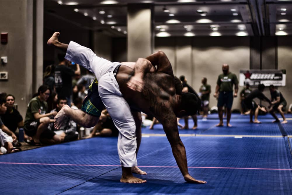 MMA fighters grappling on a training mat in a large gym while other students look on.