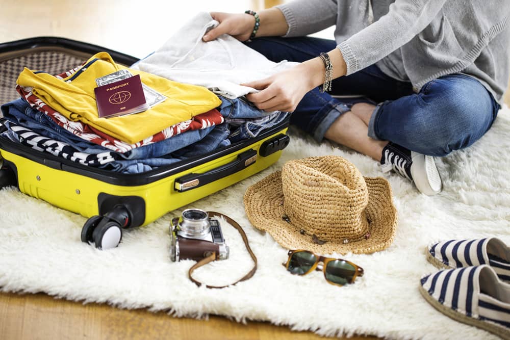 Traveler in jeans and a sweater packing a yellow suitcase with denim, a sunhat, passport, and camera.