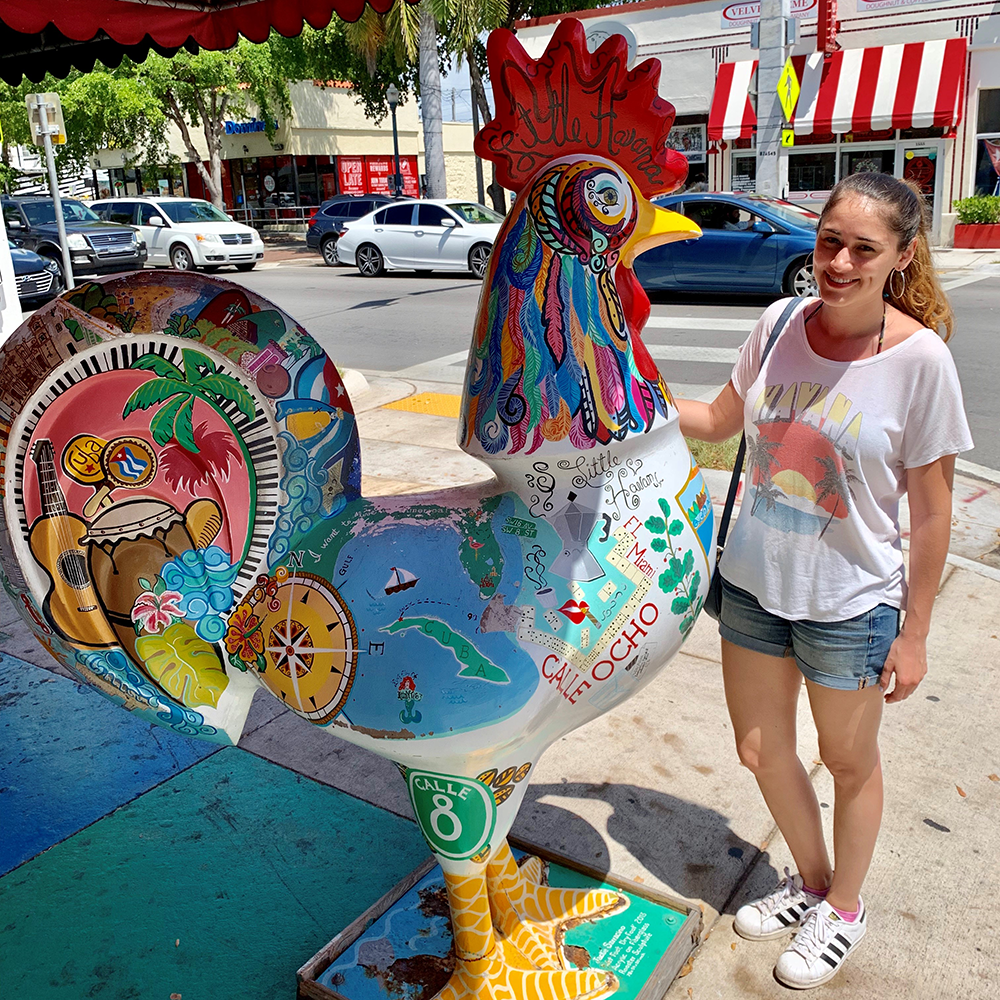 Veronica stands next to an elaborated painted statue of a rooster on Calle Ocho in Little Havana, Miami.