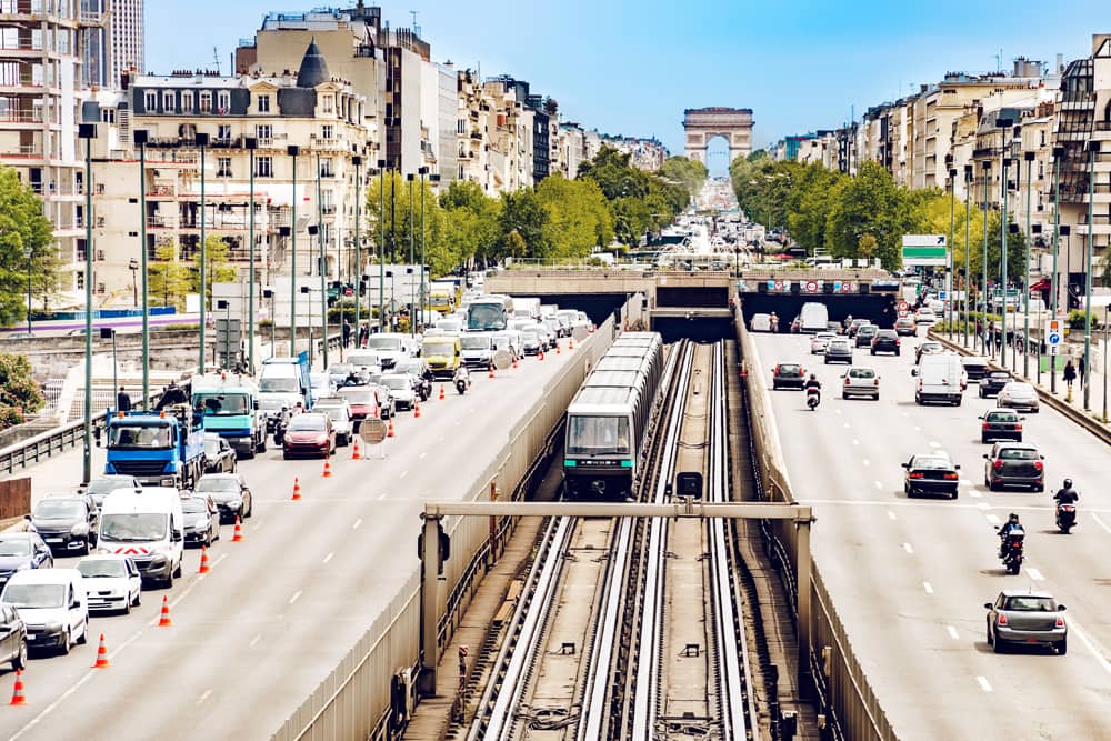 Different ways to get around Los Angeles during LA Food and Wine Festival. A Metro train on the tracks with four lanes of freeway on both sides. The sky is clear and blue, and there are several cars on the road, but traffic is light.