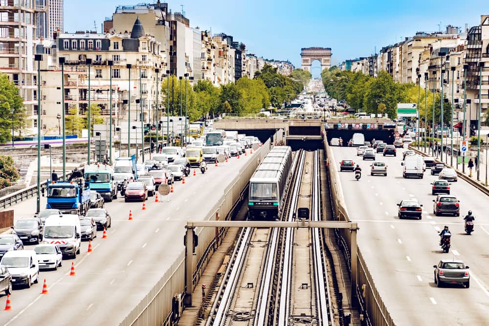 A Metro train on the tracks with four lanes of freeway on both sides. The sky is clear and blue, and there are several cars on the road, but traffic is light.