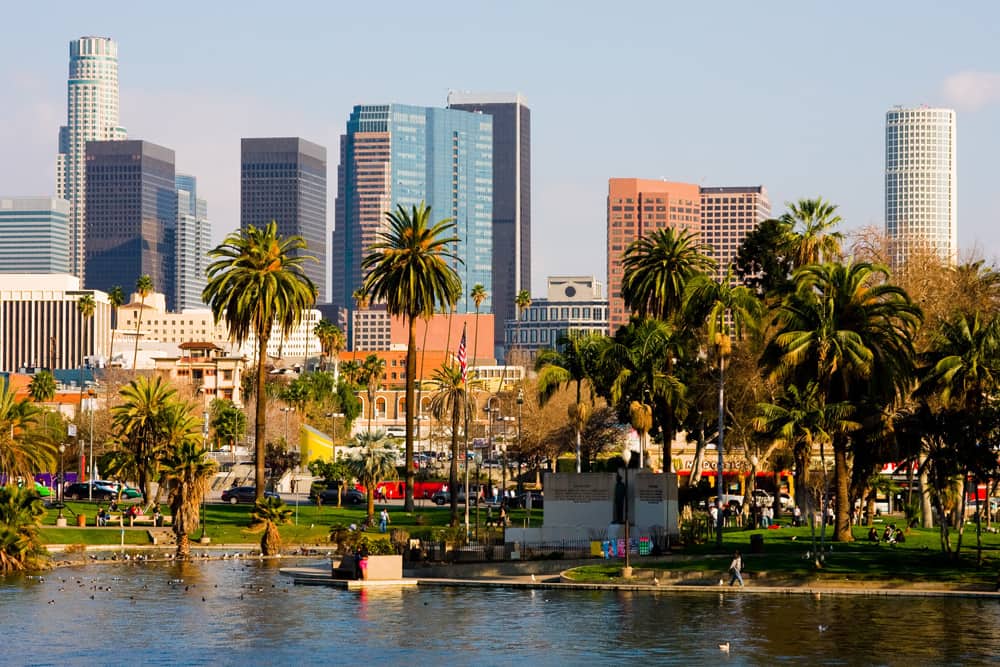 Skyline of downtown Los Angeles behind the waterfront of a lake with several palm trees and families. It is a sunny afternoon with a clear sky.