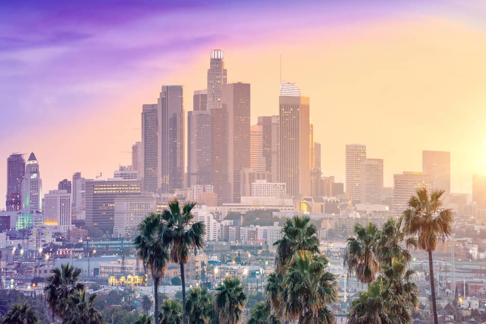 Skyline of downtown Los Angeles high-rises in from a distance at sunset. The lights are on in some of the windows and the sky is purple and yellow.