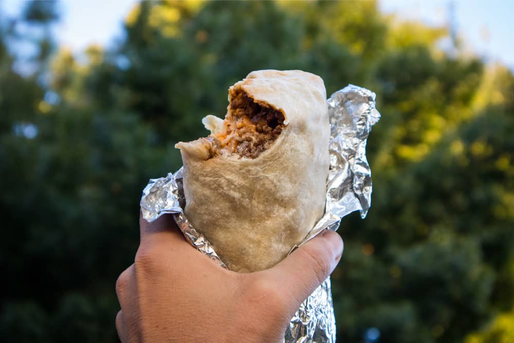 Hand holding up a burrito, revealing meat, cheese, and sour cream—ingredients in a classic Mission-style burrito.