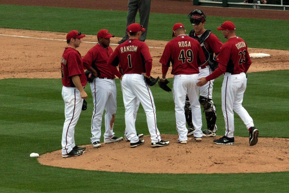 Members of the Cactus League talking on the pitcher's mound in their red and white jerseys.