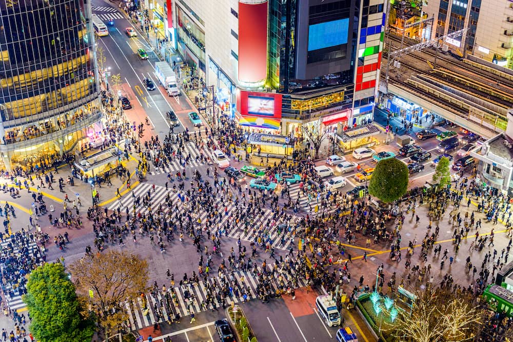 The famous Shibuya crossing in Tokyo, Japan. Hundreds of pedestrians cross the busy intersection at night. Buildings and neon signs surround them.