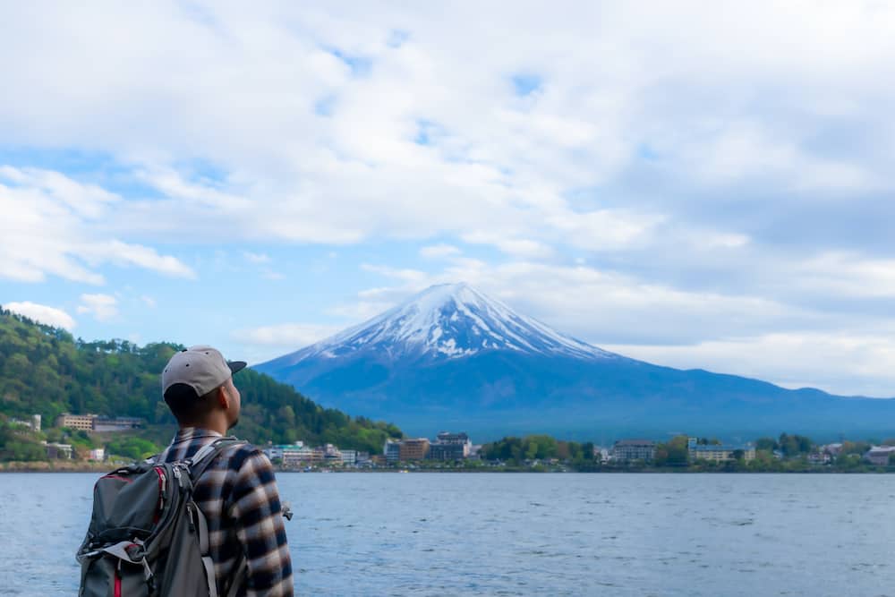 A man wearing a backpack stands in front of a lake and gazes at Mt. Fuji in the distance.