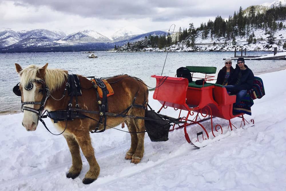 A horse-drawn carriage with two people enjoy a ride in the snow on the banks of Lake Tahoe, which is a top place to travel to for Christmas.