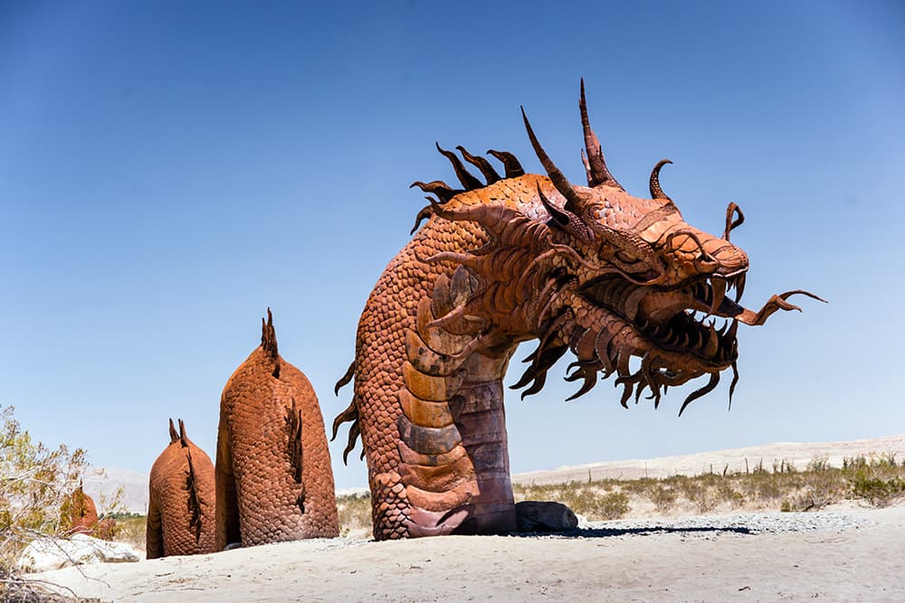 Giant red bronze statue of a dragon with pronged tongue appears to be swimming in desert sands