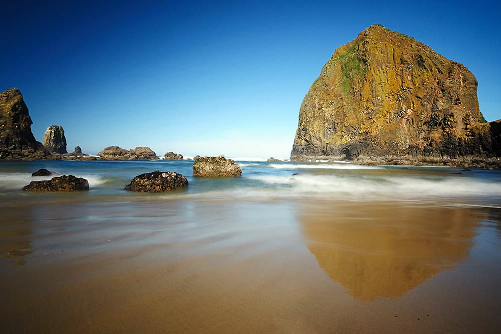 giant Haystack Rock reflected in ocean waves receding on a sandy beach