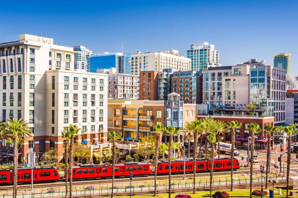 The red trolley passes palm trees and historical and high-rise buildings in downtown San Diego on a sunny day.